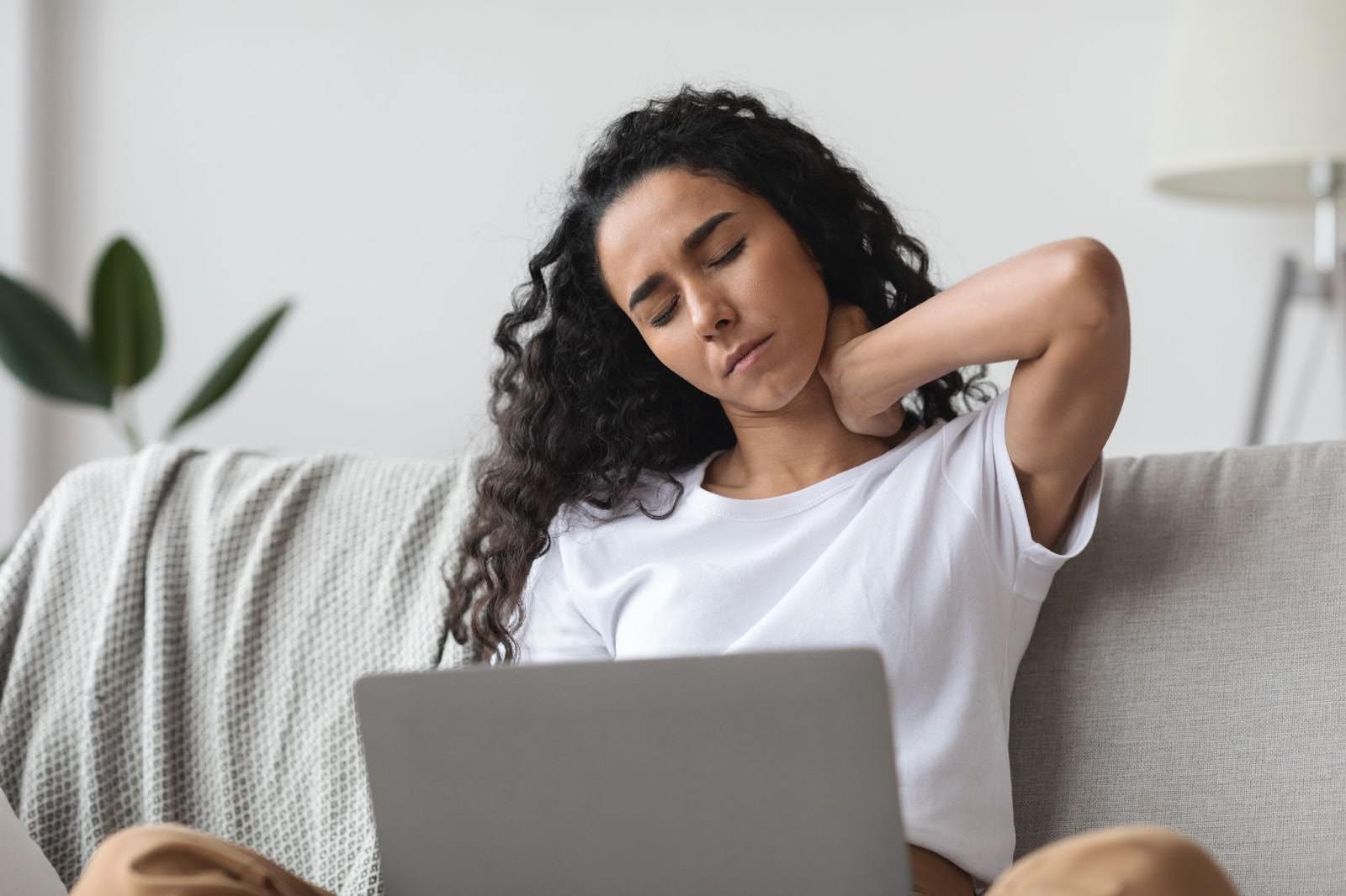 Woman sitting on a couch using a laptop, holding her neck in discomfort from pain or strain.