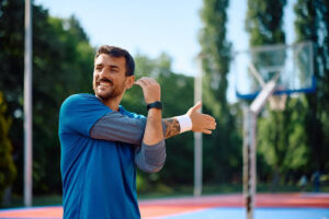 Man stands on basketball court performing arm stretches.